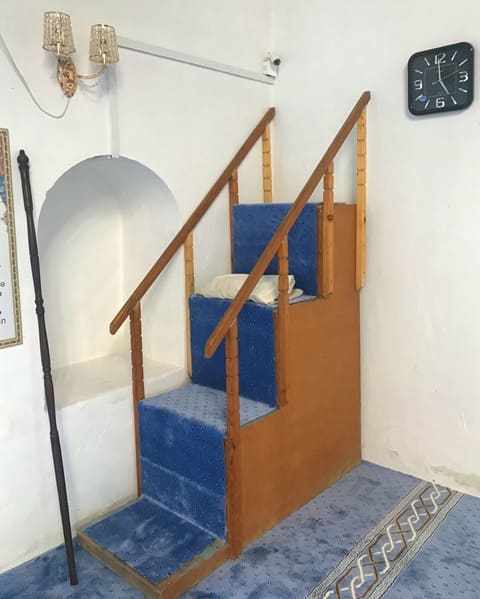 Nogai Ishan Mosque. A minbar (pulpit) for delivering sermons, often found in mosques. The structure is made of light wood with blue carpeting on the steps. It is equipped with simple wooden railings. 2024