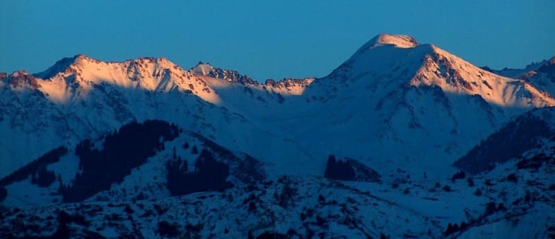 The snow-capped mountain peaks of the Trans-Ili Alatau range near Almaty, illuminated by the sun's rays against the blue evening sky.