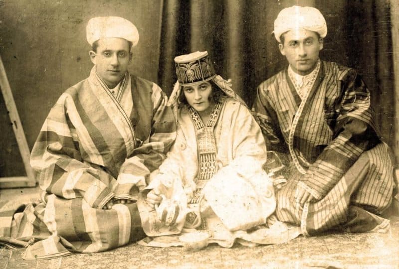На фотографии запечатлен момент из жизни бухарских евреев в Центральной Азии. Члены семьи в традиционных костюмах, характерных для начала XX века. Мужчины одеты в полосатые халаты (парча или шелк) и тюбетейки, а женщина - в праздничном платье с головным убором. Бухарские евреи исторически проживали в Центральной Азии (в основном на территории современного Узбекистана и Таджикистана), сохраняя уникальную культуру, сочетающую еврейские традиции и местные обычаи. 