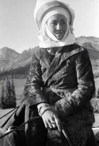 A young woman in traditional elechek costume and headdress, riding a horse, against a mountain backdrop. The photograph reflects the culture and everyday life of the peoples of Central Asia in the first half of the 20th century. The photograph is part of a series by renowned Soviet photojournalist Max Alpert, created in the 1930s. The woman is dressed in a traditional outer garment and an elaborate white headdress, typical of traditional women's costume in the region during that period.